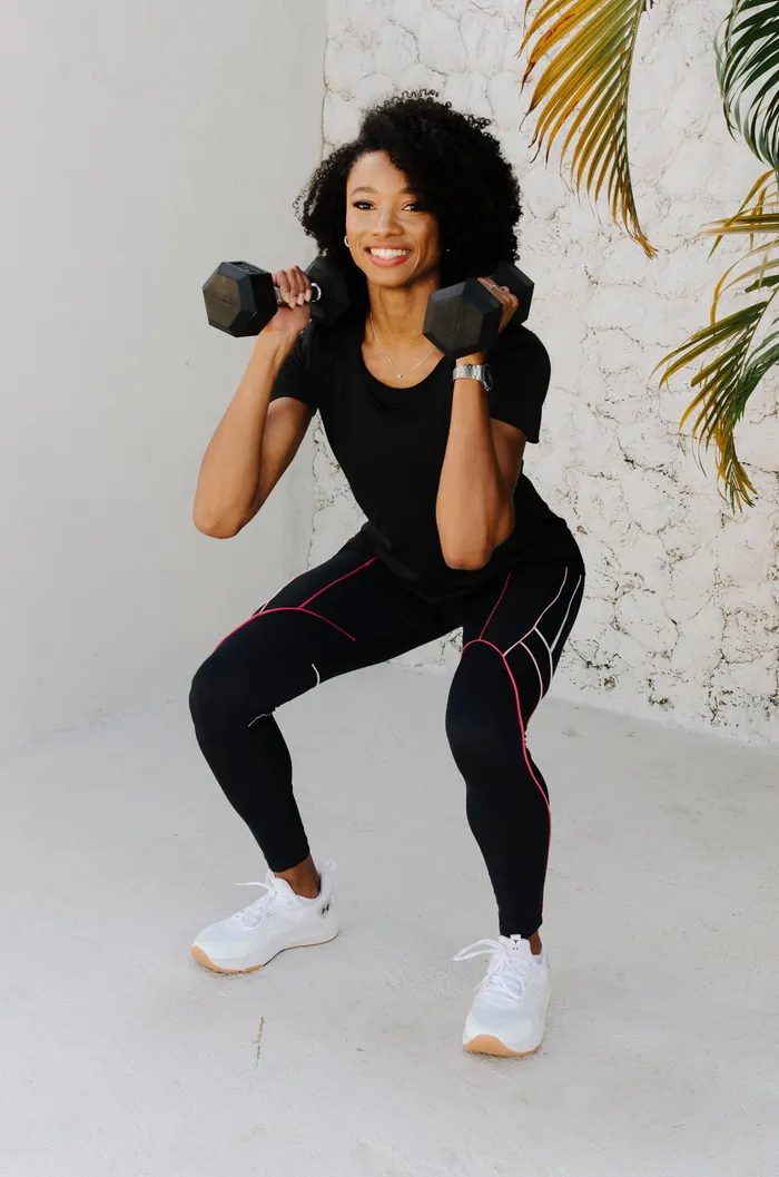 Gabby sorrindo, agachada com um halter em cada ombro